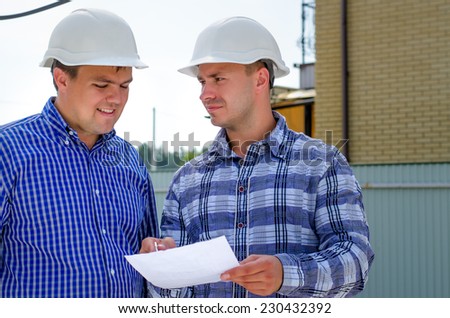 Two builders or engineers in their hardhats standing discussing paperwork and making notes outdoors on a building site