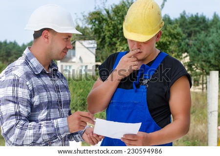Architect and builder looking at paperwork together on the building site with the builder looking perplexed with his hand to his chin