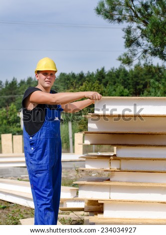Builder selecting an insulated wooden wall panel from a large stack of supplies on the outdoors building site giving the camera a quizzical serious look