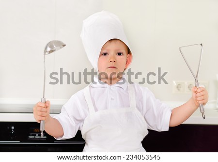 Proud little boy chef in a white toque and apron holding up a stainless steel kitchen ladle and potato masher as he stands in the kitchen