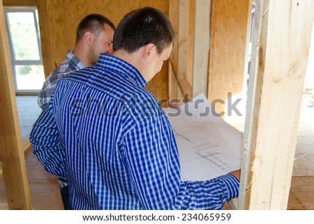 Two carpenters or builders discussing a plan of the incomplete interior of a timber frame house during the construction process, over the shoulder view of the blueprint