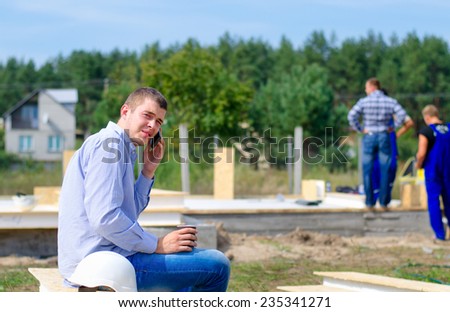 Young builder chatting on his mobile during a coffee break as he relaxes sitting on building material on the site while the team continue working in the background