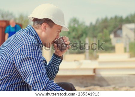 Architect taking a welcome coffee break sitting in his hardhat sipping coffee from a takeaway plastic mug on the building site, side view