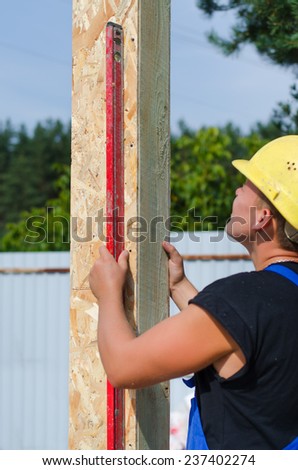 Builder ensuring that a beam is vertical holding a builders level against the wooden panel as he installs it on a new building during construction