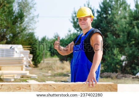 Young builder in his hardhat and overalls standing holding an insulated wooden wall panel giving a thumbs up gesture of approval and success to show all is going well