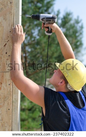 Builder or carpenter drilling a hole with a handheld electric drill in an upright wooden beam on a building site, close up side view