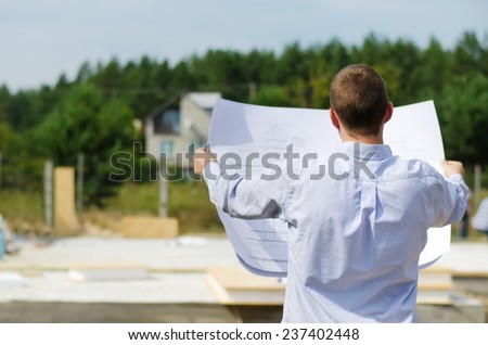 View from behind of a young architect or engineer checking a building plan on site holding it open in his hands as workmen work in the background