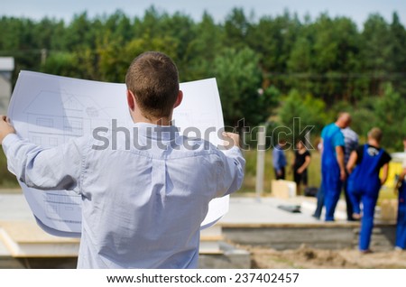 View from behind of a young architect or engineer checking a building plan on site holding it open in his hands as workmen work in the background