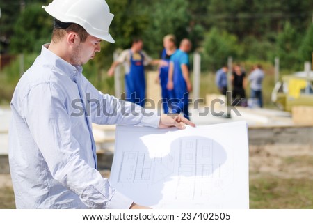 Architect or engineer checking plans on site holding the open blueprint in his hands as builders work in the background