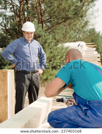 Architect or foreman watching a builder drilling a hole in a large wooden beam, view over the workmans shoulder