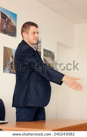 Young businessman standing over his desk offering his hand to finalise a business deal or agreement or begin a new partnership