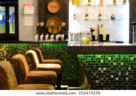 Empty bar with a tumbler of whiskey on the counter in front of a row of empty seats with the beer taps and bottles on shelves in the background