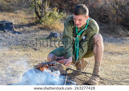 Boy Scout in Uniform Crouching and Cooking Sausages on Sticks over Campfire