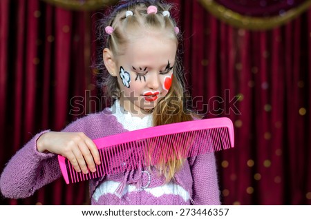 Young Blond Girl with Face Painted in Clown Make Up Brushing Hair with Over Sized Pink Comb while Standing on Stage with Red Curtain in Background