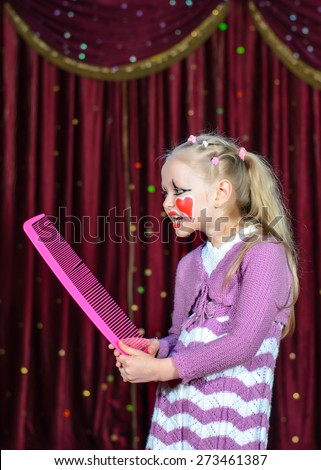 Young Blond Girl Smiling with Face Painted in Clown Make Up Holding Bright Pink Over Sized Comb on Stage with Red Curtain