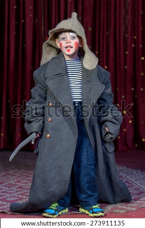 Young Boy Dressed in Over Sized Grey Coat and Floppy Hat Holding Prop Toy Sword with Face Painted in Clown Make Up, Standing on Stage Looking at Camera