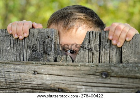 Close up Male Young Kid Peeking Over a Rustic Wooden Fence While Holding the Edge and Staring at the Camera