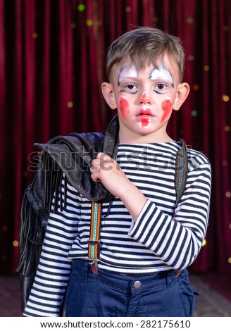 Head and Shoulders of Young Boy Wearing Clown Make Up Holding Leather Jacket Over Shoulder and Looking Solemnly Downward on Stage with Red Curtain