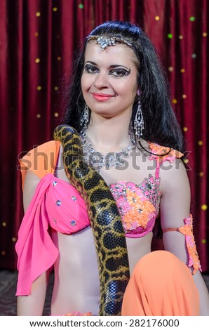 Exotic Female Dance Performer in Brightly Colored Costume with Large Constrictor Snake on Stage in front of Red Curtain