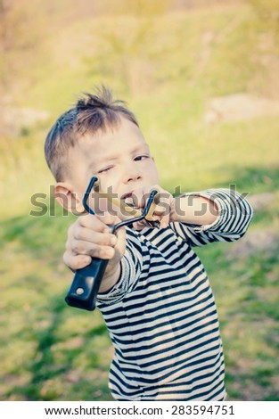 Waist Up of Young Boy Wearing Striped Shirt Holding Sling Shot with Rubber Bands Pulled Back in Anticipation of Catapulting Projectile Towards Camera