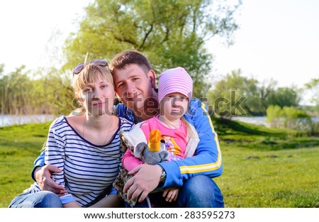 Family Portrait of Smiling Mother and Father with Pouting Young Daughter Sitting in Grass by River on Sunny Day