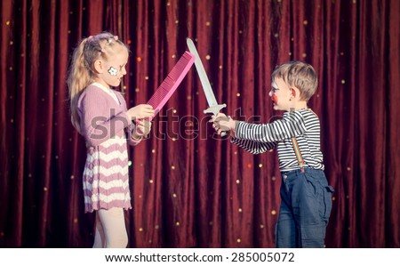 Boy and Girl Wearing Clown Make Up and Having Sword Fight with Props in front of Red Stage Curtain