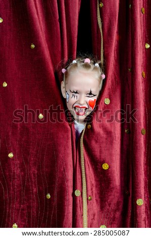 Young Blond Girl with Face Painted in Clown Make Up Smiling and Peeking Out Through Opening in Red Stage Curtains
