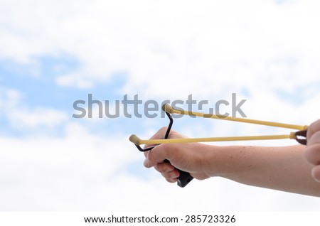 Close up Human Hand Pulling Bands of his Stone Shooter Stick Against White and Blue Sky.