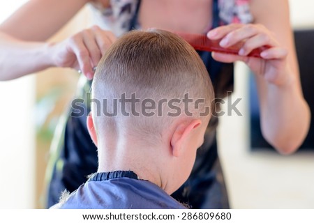 Attractive blond hairdresser cutting a young boys hair in a short hairstyle bending forwards as she trims the front, viewed from behind the child