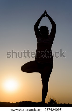 Woman practicing yoga standing balanced on one leg as she meditates in nature silhouetted at sunset by the fiery glow of the orange sun, with copyspace