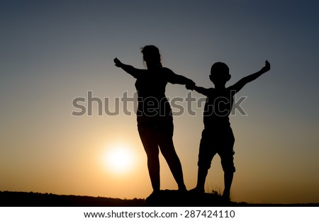 Mother and son standing holding hands on a rock with outspread arms silhouetted by the orange orb of the setting sun at dusk, with copyspace