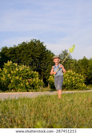 Cute funny little boy wearing hat, sleeveless top and short pants, while running to catch butterflies with a net on the green lawn in a warm day of summer