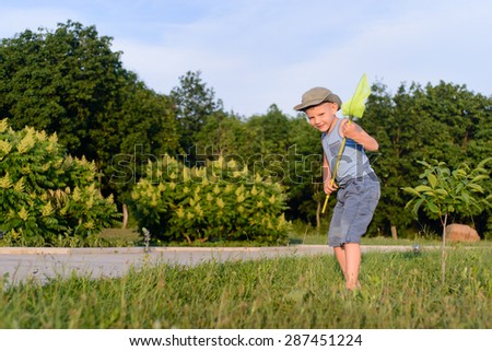 Cute funny little boy wearing hat, sleeveless top and short pants, while running to catch butterflies with a net on the green lawn in a warm day of summer