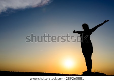 Woman embracing the sunrise or sunset standing silhouetted with outspread arms backlit by the fiery orb of the sun, spiritual image with copyspace