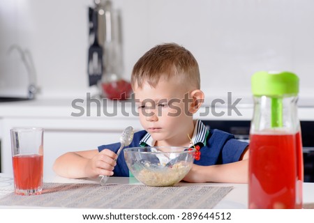 Young Blond Boy at Kitchen Table Eating Bowl of Oatmeal Cereal for Breakfast with Glass of Red Juice