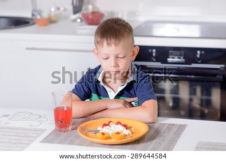 Young Unsure Blond Boy Sitting with Arms Crossed at Kitchen Table and Looking Down at Plate of Cheese and Fruit and Glass of Red Juice