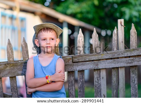Cute Young Boy Leaning Against the Wooden Fence with Arms Crossed Over his Stomach, Showing a Pensive Facial Expression.