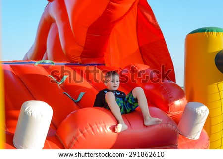 Blonde boy (7-9 years) wearing t-shirt and multi-colored shorts sitting in red bouncy castle, blue sky in background