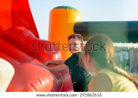 Small blonde boy (6-8 years) wearing t-shirt playing on red bouncy castle looking at camera, rear view of brunette girl (9-10 years) in foreground