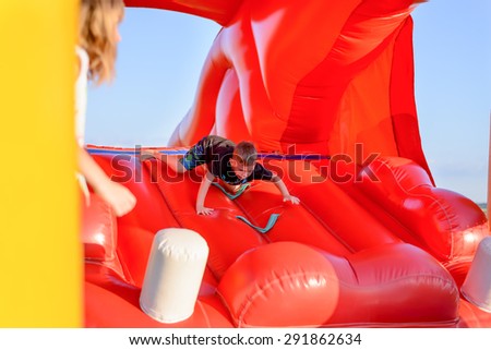 Small boy (7-9 years) wearing black t-shirt and shorts scrambles down slide of bouncy castle, part of blonde girl visible in foreground, blue sky in background