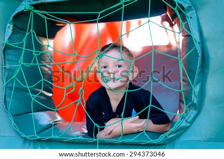 Cute little boy playing in a jumping castle peering through the net covering the window in the side grinning at the camera