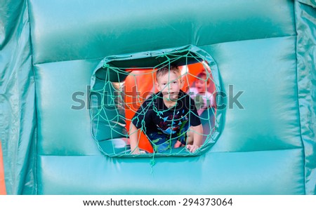 Small curious blonde boy (6-8 years) wearing t-shirt looking through netted window of blue bouncy castle, children in background