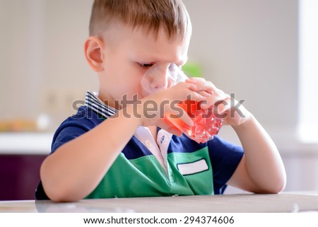 Young Blond Boy Sitting at Kitchen Table Drinking Tall Glass of Red Juice