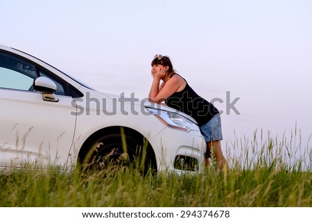 Calm Brunette Woman Leaning on Hood of White Family Car Parked in Green Field with Long Grass and Enjoying View of Sunset While Leaning on Elbows
