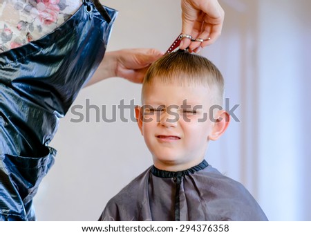 hairdresser trimming a little boys hair with a pair of scissors as her pulls a comical face with his eyes closed