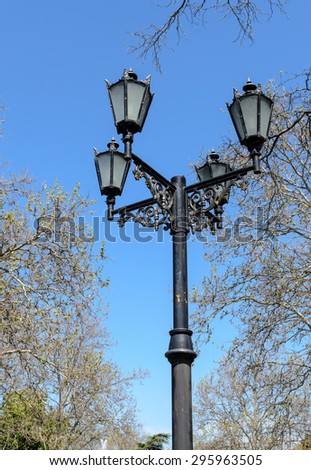 Ornate vintage wrought iron lamppost with four lanterns above intricate scroll work against a blue sky and tree viewed from below