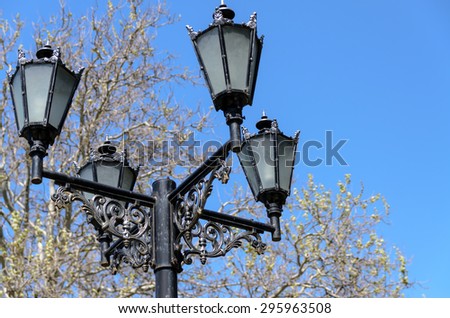 Ornate vintage wrought iron lamppost with four lanterns above intricate scroll work against a blue sky and tree viewed from below