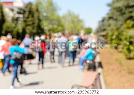 Blurred crowd of people walking along an outdoor concrete walkway between rows of trees and sitting on benches in the sunshine in a background image of a crowd scene