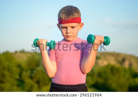 Half Body Shot of an Athletic Cute Boy with Red Warrior Headband, Lifting Two Small Dumbbells and Looking at the Camera Against Green Mountain and Sky.