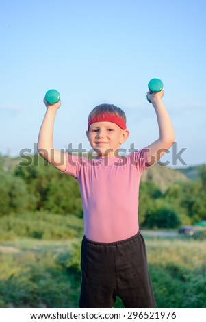 Half Body Shot of a Smiling Strong Young Boy with Warrior Headband, Raising Two Green Dumbbells and Smiling at the Camera Against Nature Background.